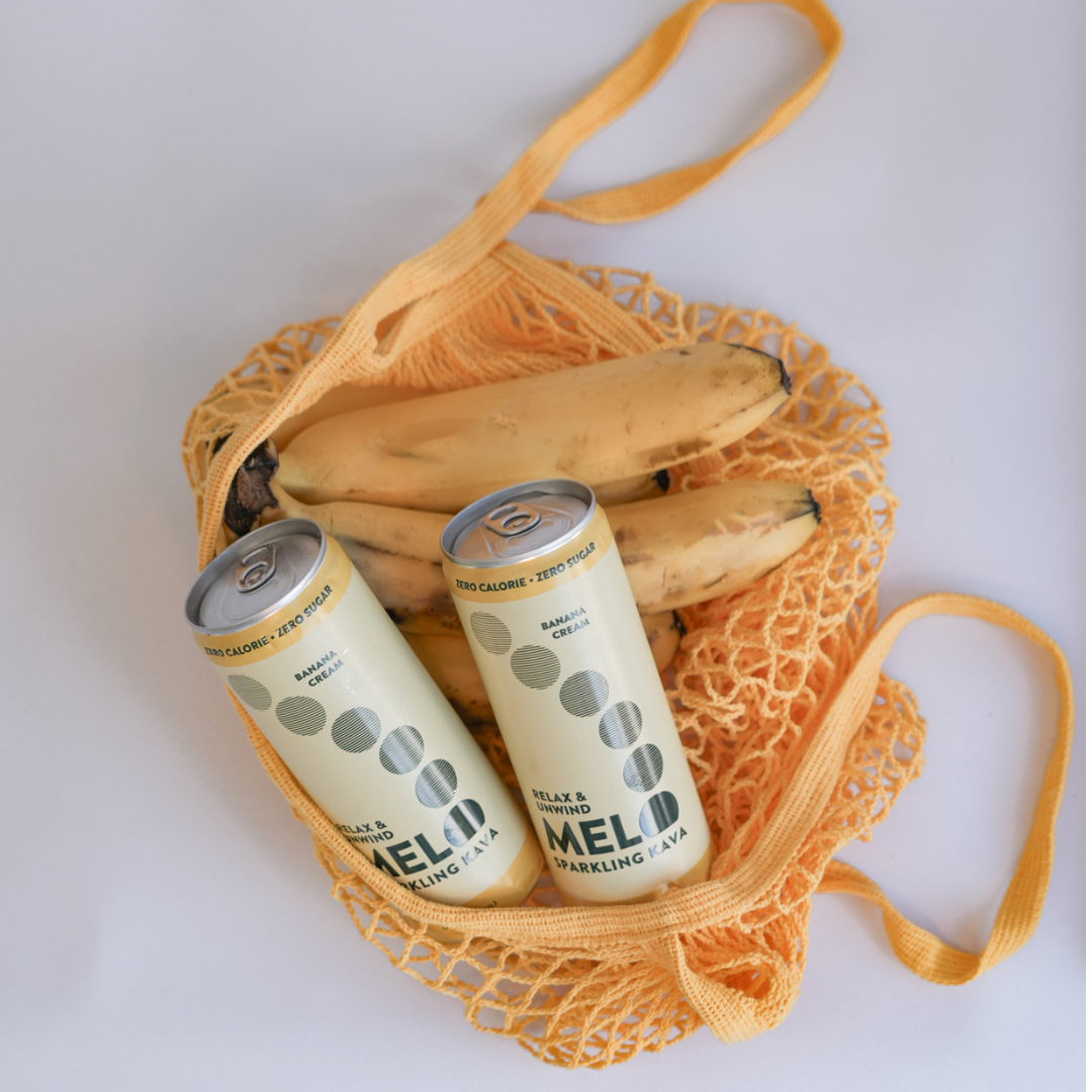 Two cans of Melo sparkling water with bananas in a yellow mesh bag on a white background