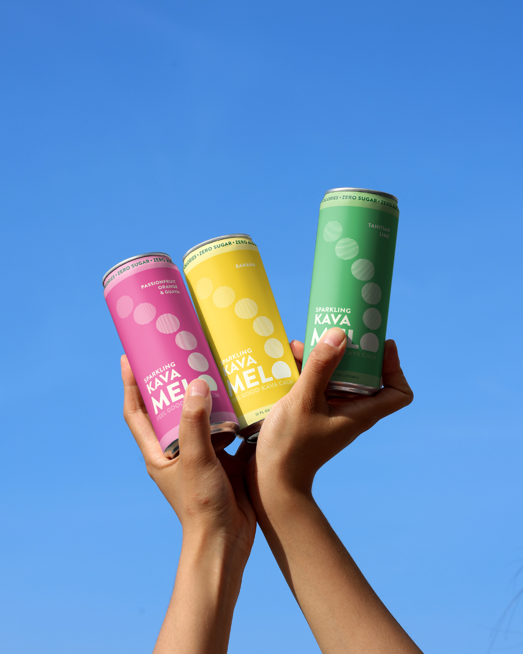 Three colorful cans of Kava Melo held against a clear blue sky.