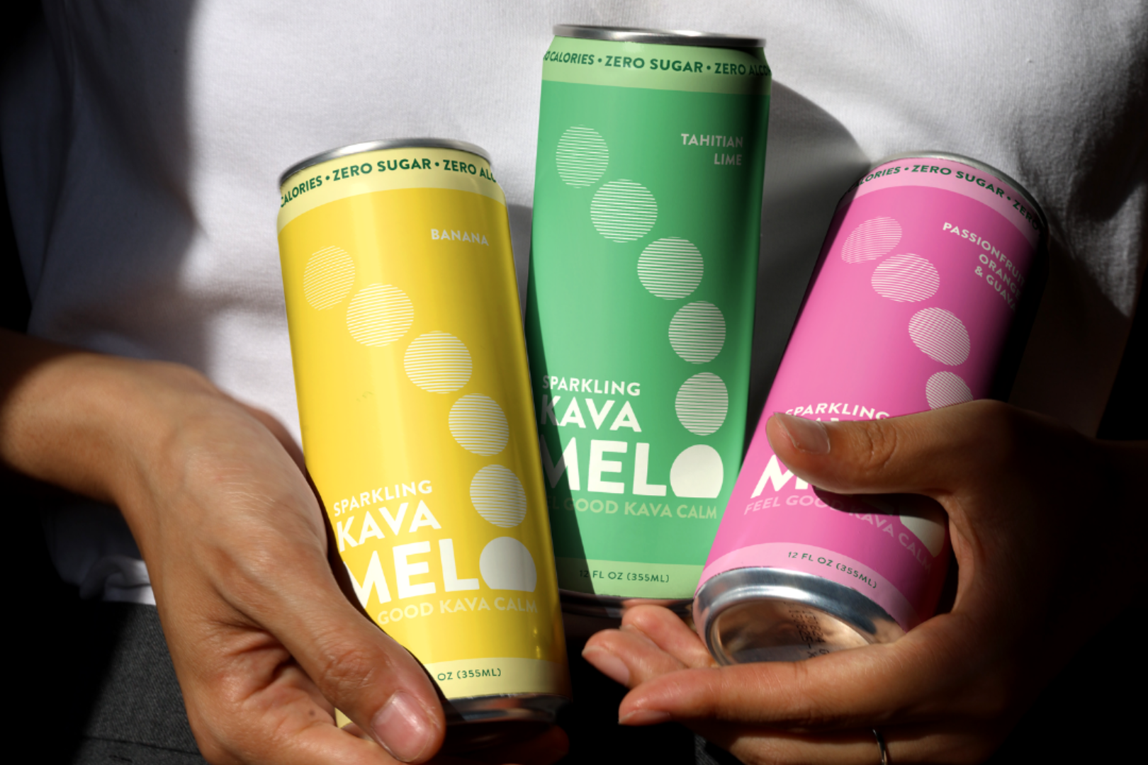 Three colorful cans of Sparkling Kava Melo held by a person against a neutral background.