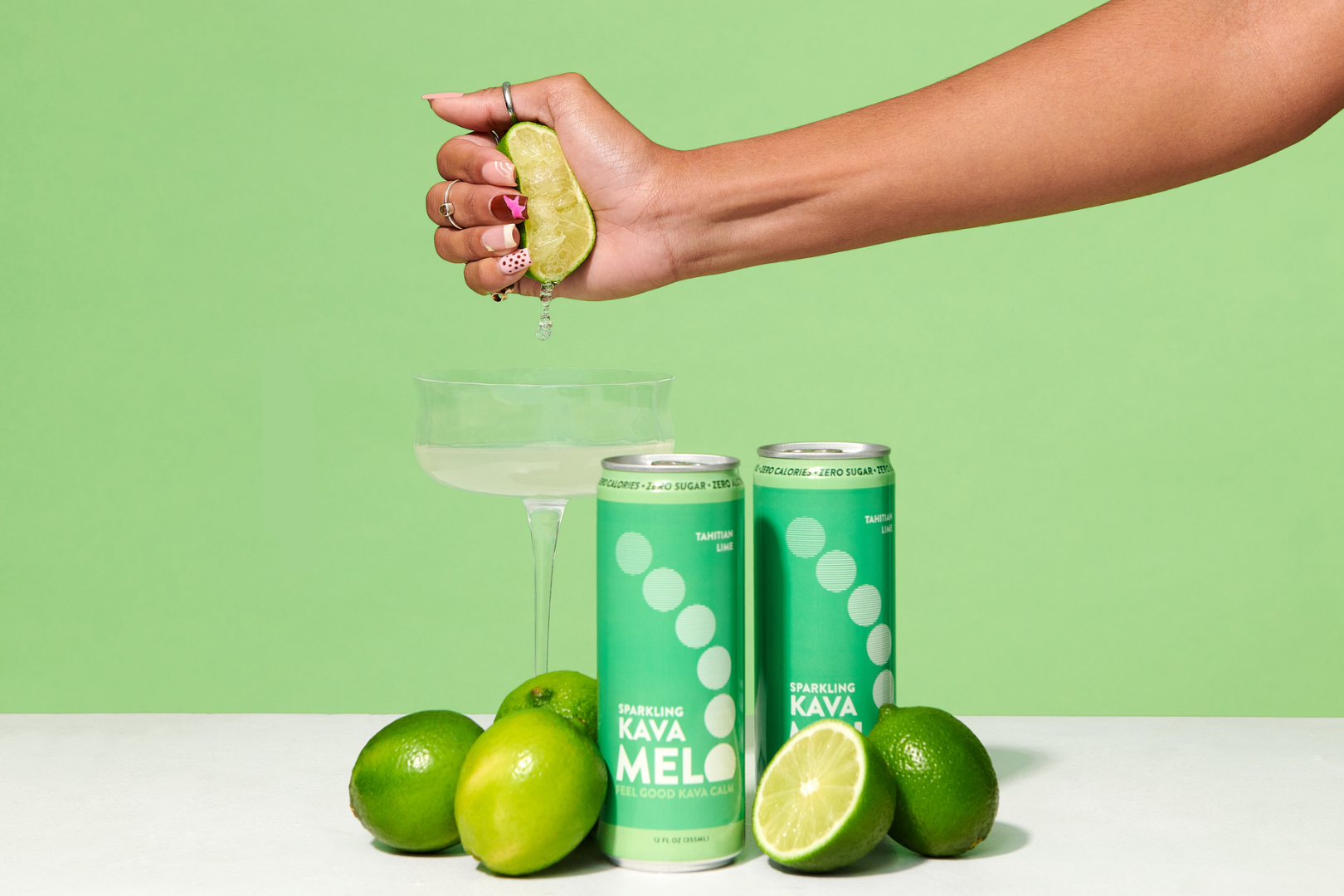 Person squeezing lime into a glass with Kava Mel cans and limes on a green background