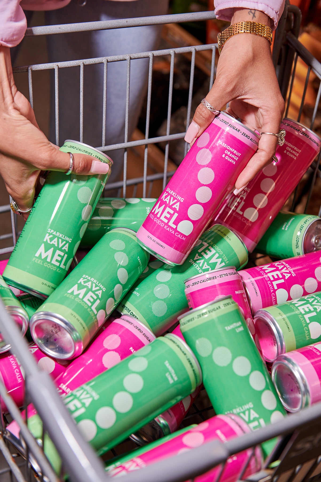 Cans of Kava Melo in green and pink being held above a shopping cart filled with more cans.