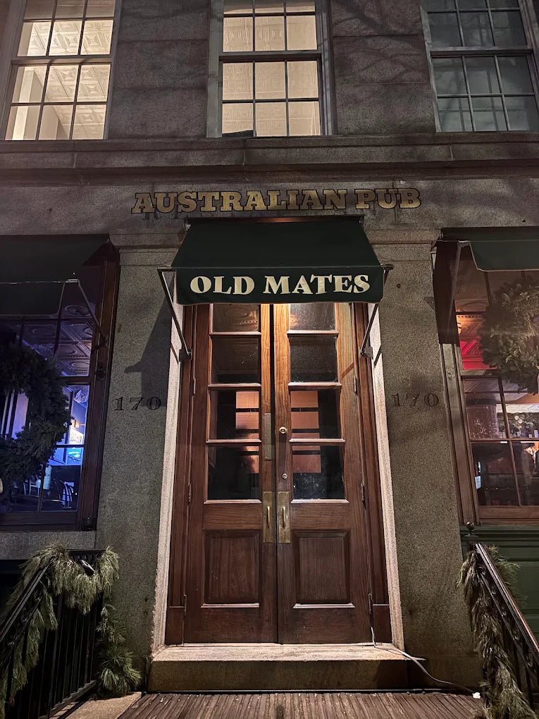 The front entrance of the Old Mates Australian Pub at night, featuring a dark green awning with white lettering, double wooden doors, and stone facade walls.