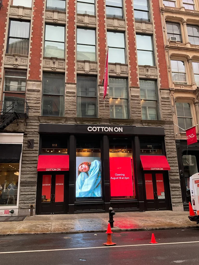 Cotton On retail storefront on a city street, with a brick and stone building facade, red awnings, large display windows, and orange traffic cones on the wet sidewalk.